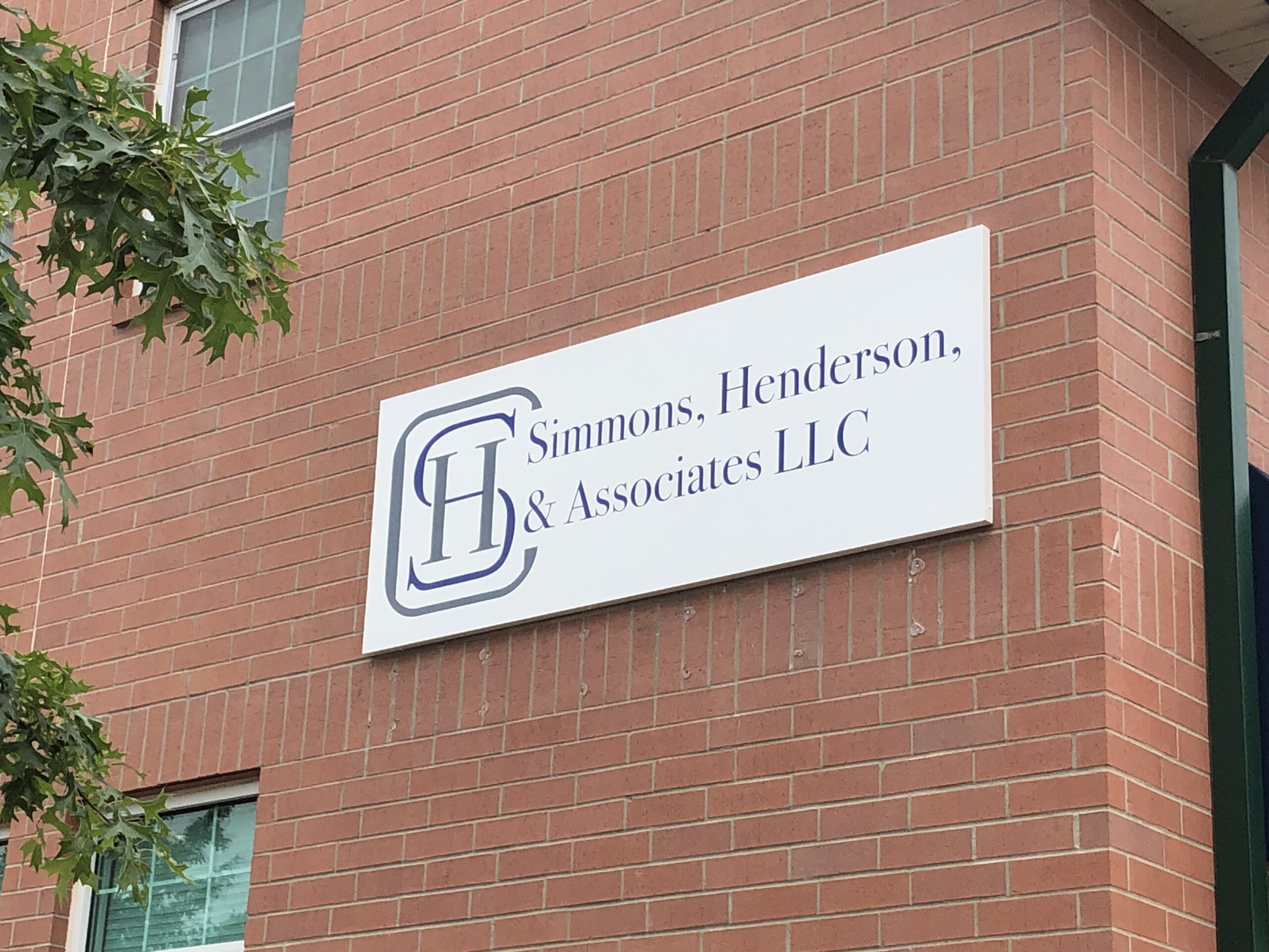 The corner of a red brick building is seen with a few green oak leaves framing the left edge. A large sign hangs on the brick showing a logo for the attorney's office.